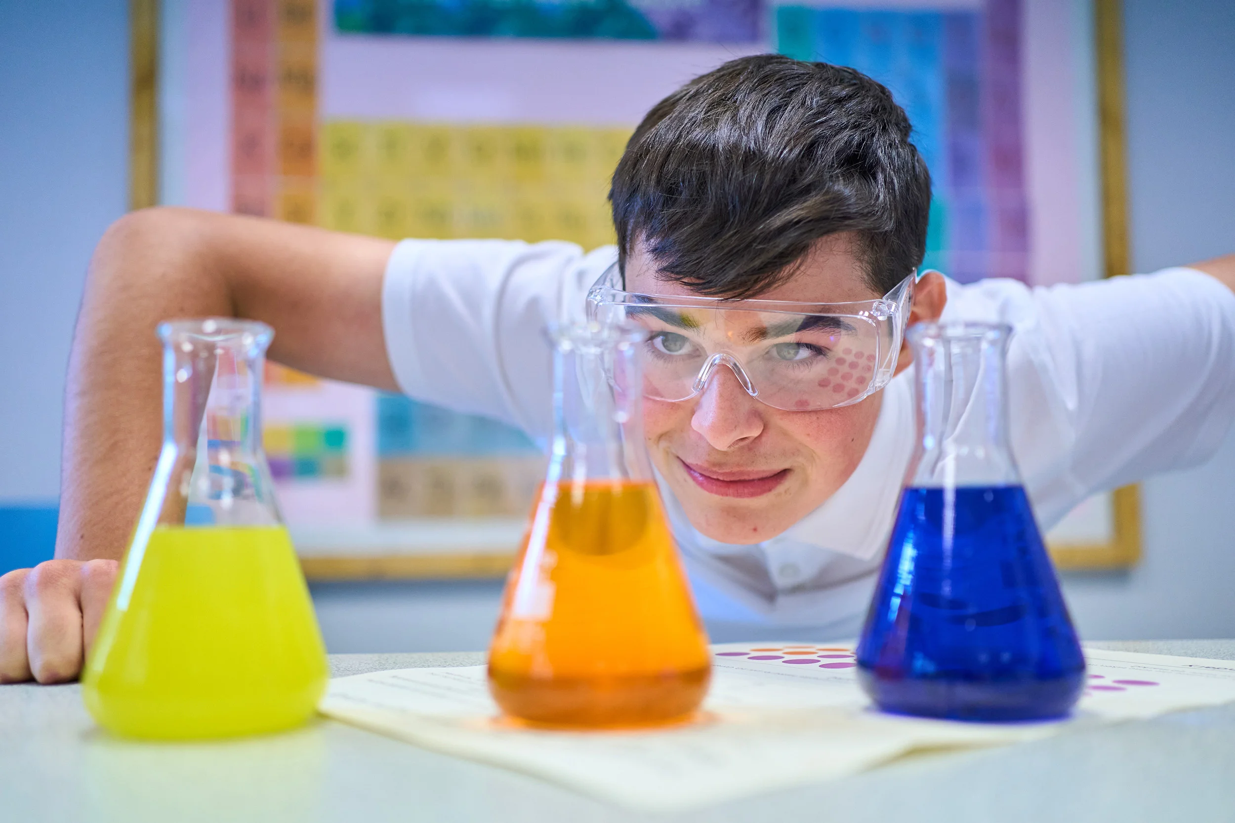 school pupil wearing safety goggles looks at coloured liquids in scientific containers